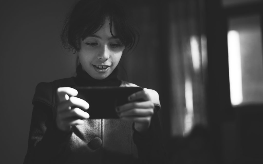 A young girl smiling while using a smartphone in a black and white setting indoors.